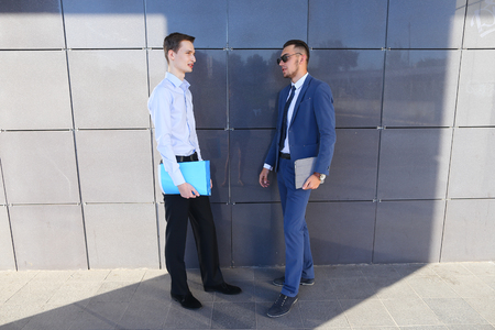 Two beautiful active young male businessman guys discussing important issues, make decisions, solve problems, share business ideas, smiling, holding in hands colored folders and stand opposite each other near gray wall of modern business center on outdoorの写真素材