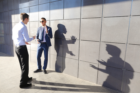 Two beautiful active young male businessman guys discussing important issues, make decisions, solve problems, share business ideas, actively gesticulating and stand opposite each other near gray wall of modern business center on outdoors. Guy with glassesの写真素材
