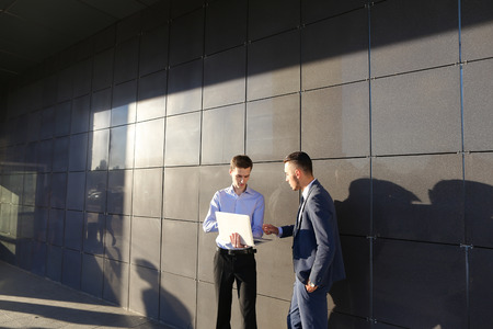 Handsome confident young male man, businessman, student holds laptop in hands and deliver presentations, talks about gadget, advertises new technology to colleague who listening to attentively, and stand on background wall of modern business center outdooの写真素材