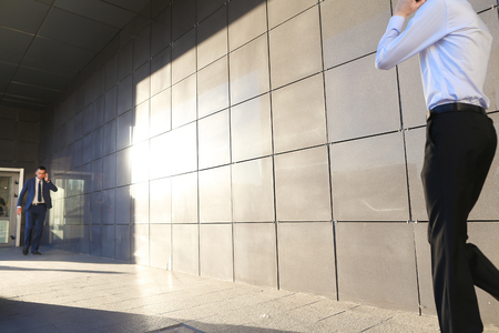 Young perspective male men, students,go to meet each other, greet one another, shake hands, talking on phone, stop and talk, walk fast step, hurry, running late on background of wall of business center outdoors .One of guys dressed in blue classic suit, wの写真素材