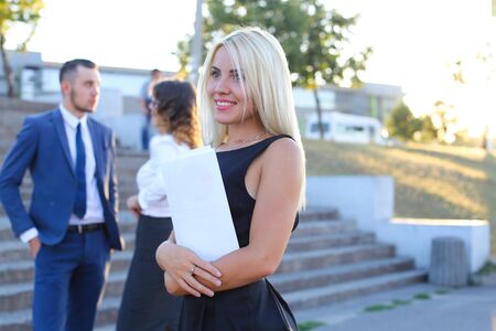 Pretty girl smiling, posing, holds paper in hands and looking away on background of people outdoors. Blonde girl dressed in black dress. Concept of business meeting, business communication, successful people, business clothing advertising, deal.の写真素材