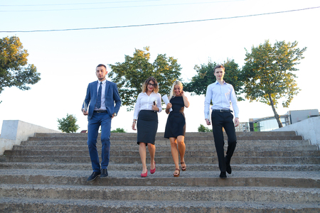 Young modern confident people, two boys and two girls, perspective entrepreneurs, students smiling, hold in hands gadgets, tablet, smartphone and papers, documents, go down stairs outdoors. One of guys dressed in blue classic suit, white shirt with tie, sの写真素材