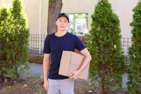 Deliveryman Carring Parcel and Giving it and Smiling. International Staffing Company Shows That Courier of Company Always Ready to Work at Any Time and Any Day, Young Man of European Appearance in Uniform, With Broad Smile and Good Mood on Background of Hの写真素材