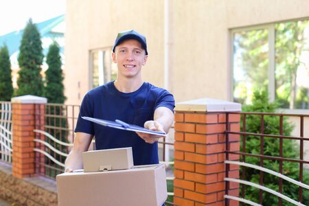 Messenger Carring Two Boxes in Hands Smiling on Background of Trees Along Street. Courier Company Provides Its Services in All Areas Ofterrain, Man of European Appearance in Uniform Carries Two Boxes With Order Along Street on Background of Shrubs, Trees の写真素材