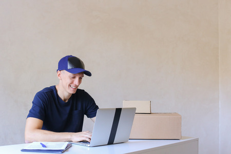 Young Postman Sits at Table Typing at Laptop. Courier Company Accepts Orders Every Day Different Types and Sizes Young Deliveryman European Appearance in Black T-Shirt and Cap Sitting at White Table Typing on Computer and Takes Pen Writing Down Informatioの写真素材
