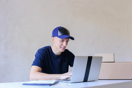 Young Postman Sits at Table Typing at Laptop. Courier Company Accepts Orders Every Day Different Types and Sizes Young Deliveryman European Appearance in Black T-Shirt and Cap Sitting at White Table Typing on Computer and Takes Pen Writing Down Informatioの写真素材