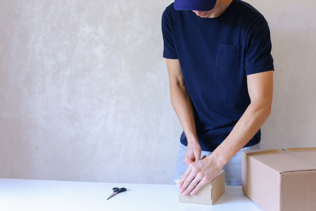 Deliveryman Standing in Room, Glue Tape Box and Takes Parcel in Hands and Smiling . Postman European Appearance in a Black T-Shirt and Cap Stands Near the Desk in the Office With White Walls and Unassisted Packaging Boxes, Adhesive Glue Tape on a Cardboarの写真素材