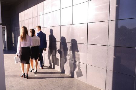 Successful male guy, businessman and two pretty young women, business people, students go in opposite direction from camera and discuss working day, returning from lunch break, study plans and kept paper in hands, graphics, and view them and go along of gの写真素材