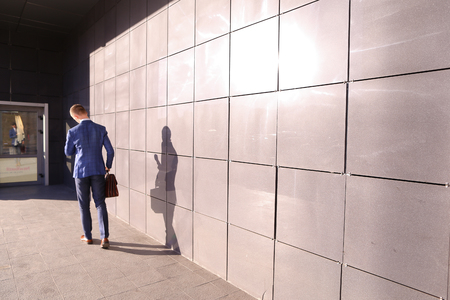 Handsome young male guy entrepreneurstudent hurry on business meeting, returns from lunch, going home after work, looks at his watch put on by on  hand, late and goes on background of gray wall of business center outdoors. Stylish man dressed in blue busiの写真素材