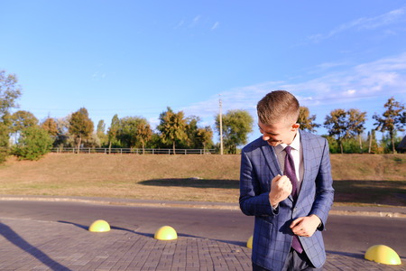Successful handsome young male man budding entrepreneurstudent stands in front of camera, posing and smiling, rejoicing success, clasps his hands in fist and shows gesture of "Yes!" on background of street and parking outdoors. Stylish man dressed in blueの写真素材
