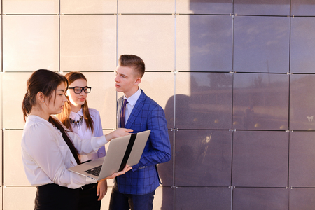 Modern young man and two attractive smart young women, business people, students communicate, holding laptop and study work plan, familiarize with presentation, teach each other new programs, solve important problems and workloads holding folder with docuの写真素材