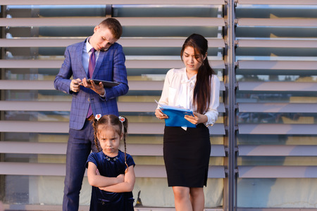 In  foreground stands and frowns, angry with arms crossed in front of little girl, child,  in background successful young handsome mans man, young father and modern woman, young mother hold gadgets in hand, mobile phones and talking to them, solve operatiの写真素材