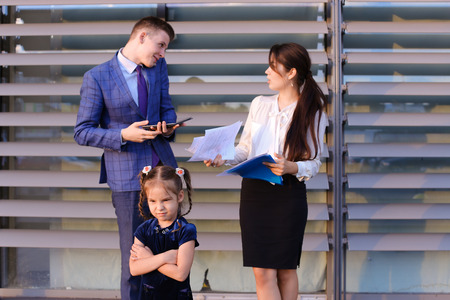 In  foreground stands and frowns, angry with arms crossed in front of little girl, child,  in background successful young handsome mans man, young father and modern woman, young mother hold gadgets in hand, mobile phones and talking to them, solve operatiの写真素材