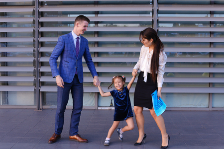 Handsome male man, young father and modern attractive girl, young mother holding hand of little girl, daughter, smile at each other, laughing and having fun, lead child in kindergarten, standing on background of large gray box business center outdoors. Stの写真素材