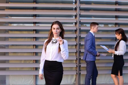 Successful young adult  business lady, student rests, took break from work, watching, smiling and posing at camera and stands on background of colleagues who discuss workflows, consider paper near of business center outside. Girls with light brown and darの写真素材