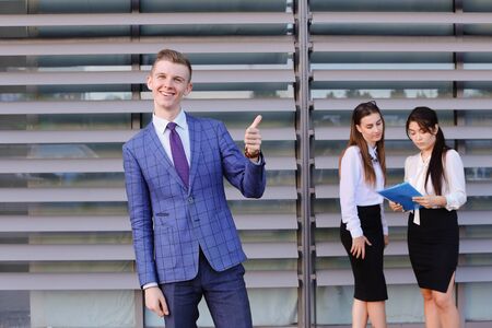 Handsome confident young male guy businessman, office worker, student posing and smiling in front of camera, shows  gesture "thumbs up" , posing for shoot of modern mens business clothing catalog and stands at colleagues background two attractive young woの写真素材