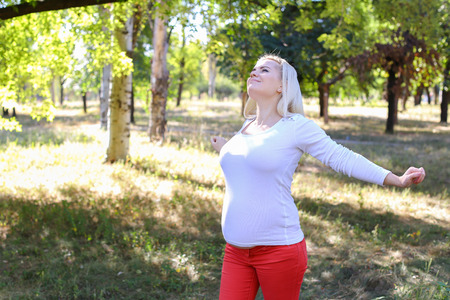 Gorgeous Young Adult Woman in Family Way of European Appearance Decided to go Outside and Get Some Fresh Air, Stretching and Warming up in Green Sunny Park Outdoors in Daytime. Female With Blonde Hair Braided in Braid in Front Dressed in White Blouse Withの写真素材