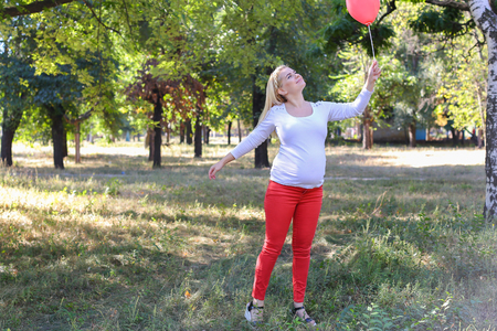 Alluring Future Mother of European Appearance Enjoys Pleasant Things and Look Forward to Appearance of First-Born Into World Photographed For Advertising of Womens Clothing For Pregnant With Pink Ballon in Shape of Heart in Green Sunny Park Outdoors in Daの写真素材