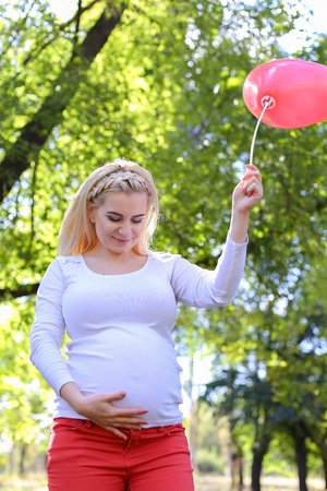 Marvelous Young Adult Lady With Child of European Appearance Enjoying Sunny Day, Cute Smiles, Poses and Rises up Pink Ballons in Shape of Heart in Green Park Outdoors in Daytime. Female With Blonde Hair Braided in Braid in Front Dressed in White Blouse Wiの写真素材