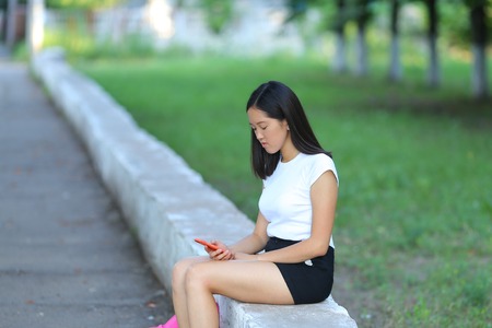 Girl female teenager with black hair in a bob white east Asian woman green park with a backpack background. Lady holds, dials, push your finger, smiling, looking into the camera, talking on the phone happy.の写真素材