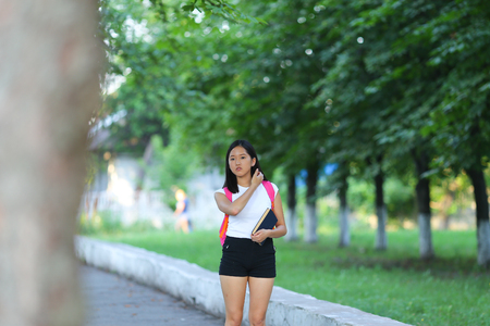 Girl female teenager with black hair in a bob white east Asian woman green park with a backpack background. Lady walking, goes, walk away, look at the camera.の写真素材