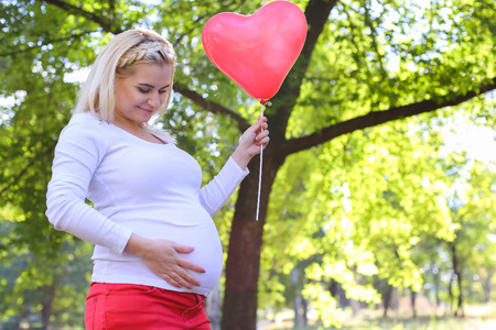 Marvelous Young Adult Lady With Child of European Appearance Enjoying Sunny Day, Cute Smiles, Poses and Rises up Pink Ballons in Shape of Heart in Green Park Outdoors in Daytime. Female With Blonde Hair Braided in Braid in Front Dressed in White Blouse Wiの写真素材