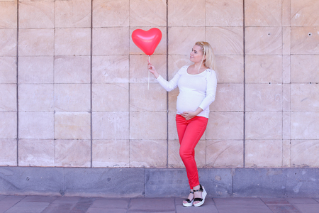 Stunning Young Adult Girl in Family Way Gently Stroking Belly in full growth on Background of Stone Wall in Daytime and Wind Develops Blond Hair. Female to Carry Child Under Ones Heart and Photographed For Social Program Mothers Day and Holding Pink Balloの写真素材