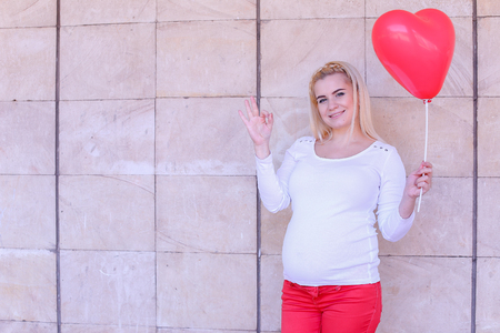 Woman to be Expecting With Blond Hair Feels Happy and Prepares to Become Mother, Holding in Hand Pink Balloon in Shape of Heart, Smiling and Posing at Camera Shows Gesture Okay. Female Stands on Background of Stone Slabs in Open Air in Daytime. Girl With の写真素材