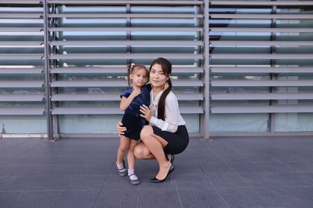 Charming modern female woman, young mother, successful business woman squatted down next to daughter, smile at each other, hug with girl, small child hugging her mothers neck and kisses on cheek, posing for camera, girl holding baby girl on background of の写真素材