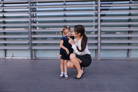 Charming modern female woman, young mother, successful business woman squatted down next to daughter, smile at each other, hug with girl, small child hugging her mothers neck and kisses on cheek, posing for camera, girl holding baby girl on background of の写真素材