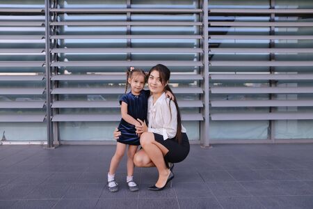 Charming modern female woman, young mother, successful business woman squatted down next to daughter, smile at each other, hug with girl, small child hugging her mothers neck and kisses on cheek, posing for camera, girl holding baby girl on background of の写真素材
