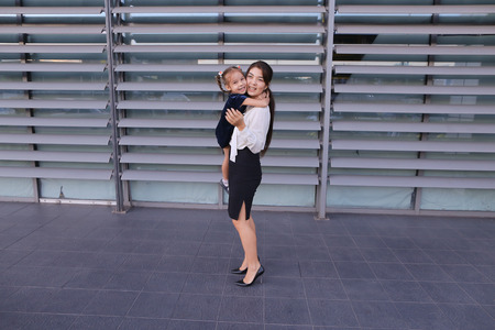 Charming modern female woman, young mother, successful business woman squatted down next to daughter, smile at each other, hug with girl, small child hugging her mothers neck and kisses on cheek, posing for camera, girl holding baby girl on background of の写真素材