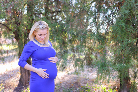 Marvelous Young Adult Female to Carry Child Under Ones Heart, Relax and Enjoy Nature and Fresh Air, and Stands in Big Green Park Outdoors in Daytime. Girl With Blond Hair, Braided in Braid in Front Dressed in Long Blue Dress. Concept of Happy Pregnancy anの写真素材