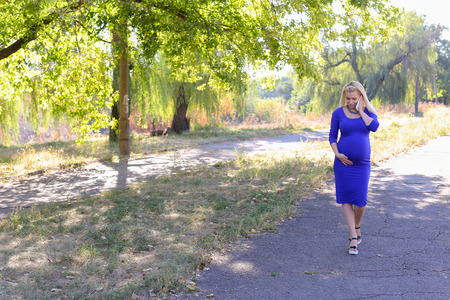 Dazzling Contemporary Young Adult Feminine of European Appearance Carries Baby Under Heart. Girl Leisurely Walking and Chatters on Phone With Husband About Well-Being and Appoint Meeting at Cinema or Says Family Doctor About State of Health. Female With Bの写真素材