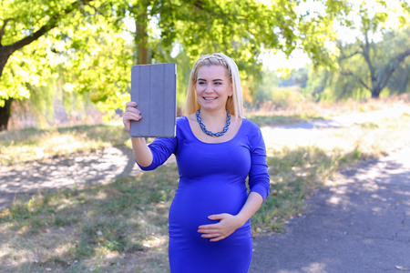 Gorgeous Contemporary Young Feminine Carries Baby Under Heart. Girl Chatters on Skype With Husband About Well-Being or Informs Parents About Anticipated Date of Childbirth. Female With Blond Braided Plait Hair in Front Dressed in Long Blue Dress With Sleeの写真素材