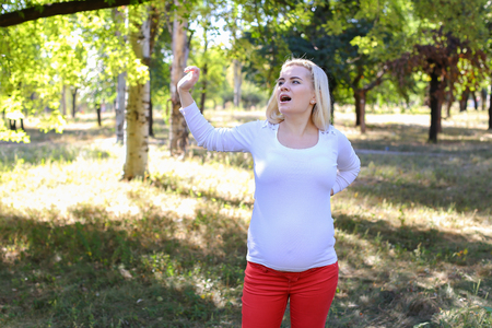 Gorgeous Young Adult Woman in Family Way of European Appearance Decided to go Outside and Get Some Fresh Air, Stretching and Warming up in Green Sunny Park Outdoors in Daytime. Female With Blonde Hair Braided in Braid in Front Dressed in White Blouse Withの写真素材