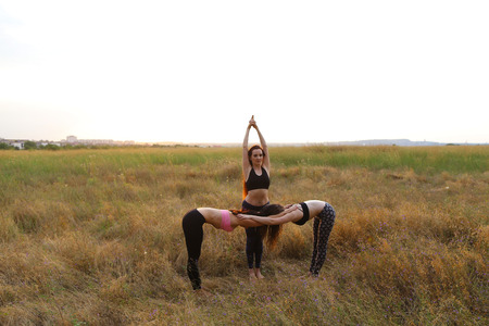 Girls perform postures or asanas of yoga, posing for photographer to take advertising pictures for sports womens clothing catalog, meditate outdoors in wide field. Girl with shoulder-length hair dressed in pink top and black leggings, girl with long red hの写真素材
