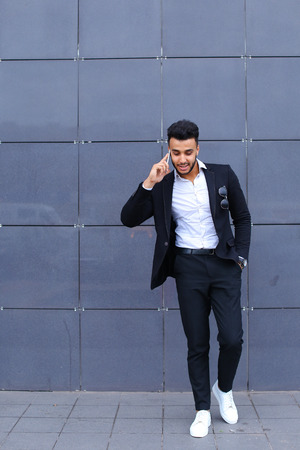 Entrepreneur man male looking at camera and smiling talks on mobile smart phone. Young handsome businessman arab muslims in business center wearing dressed in black elegant suit on building background.の写真素材