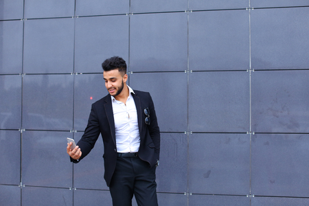 Entrepreneur takes makes selfie man male looking at camera and smiling talks on mobile smart phone. Young handsome businessman arab muslims in business center wearing dressed in black elegant suit on building background.の写真素材