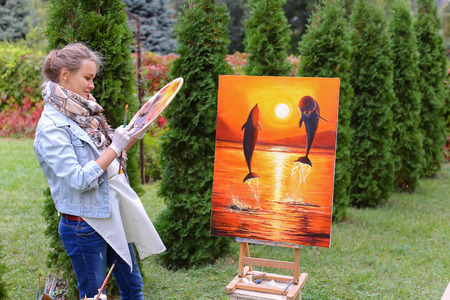 Pretty attractive artist finishes painting which depicts dolphins jumping out of  water on orange background, stands with back to photographer and behind wooden easel, holds palette smeared in paint in large green park day outdoors.Girl dressed in red sweの写真素材