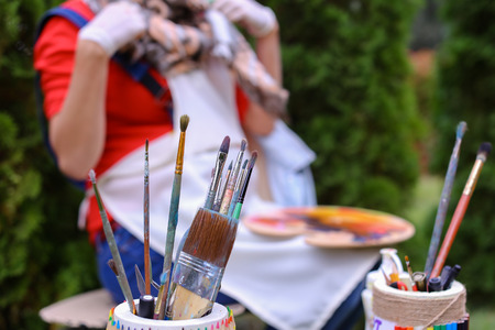 Photo shows thin and thick brush for oil and acrylic paints and pencils that stands with other artistic objects in colored jar and that lying on gray table in park in open air against girl artist. Concept of art materials store advertising art tools and pの写真素材