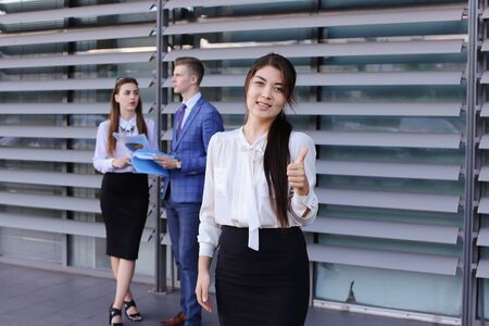 Attractive young perspective business woman, student rests, took break from work, watching, smiling and posing at camera, enjoys success, shows gesture of thumbs up, stands on background of colleagues who discuss workflows, consider paper near of businessの写真素材