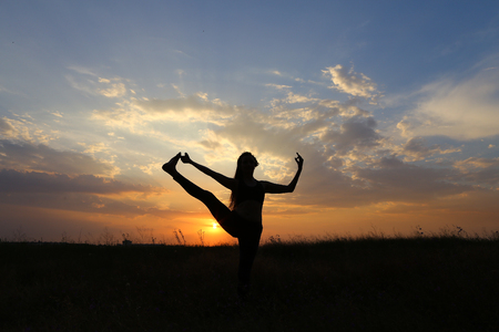 Female with beautiful figure takes yoga postures, gymnastics and ballet spends stretching and meditating, posing for advertising shoot fitness studio, healthy energy drink, dancing belly dancing among wide field on background of yellow sunset and beautifuの写真素材