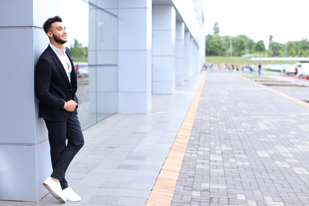Young handsome businessman Arab Muslims is in the business center in black elegant dress on building background. Businessman man male looking at time walking goes and smiling.の写真素材
