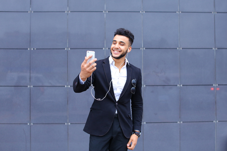 Entrepreneur takes makes selfie man male looking at camera and smiling talks on mobile smart phone. Young handsome businessman arab muslims in business center wearing dressed in black elegant suit on building background.の写真素材
