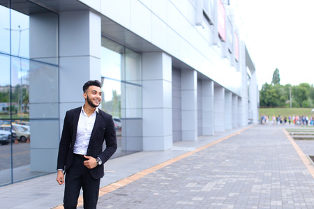 Young handsome businessman Arab Muslims is in the business center in black elegant dress on building background. Businessman man male looking at time walking goes and smiling.の写真素材