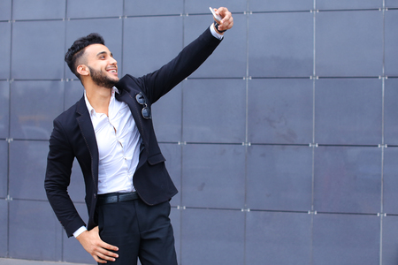 Entrepreneur takes makes selfie man male looking at camera and smiling talks on mobile smart phone. Young handsome businessman arab muslims in business center wearing dressed in black elegant suit on building background.の写真素材