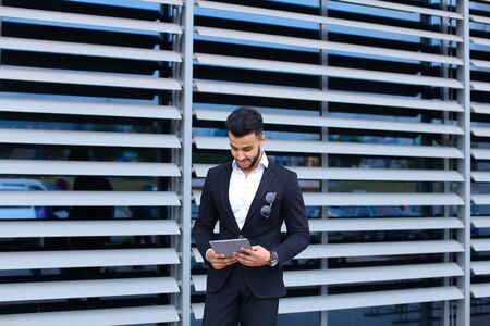 Young handsome businessman Arab Muslims is in the business center in black elegant dress on building background. Businessman man male looking waiting concept and smiling.の写真素材
