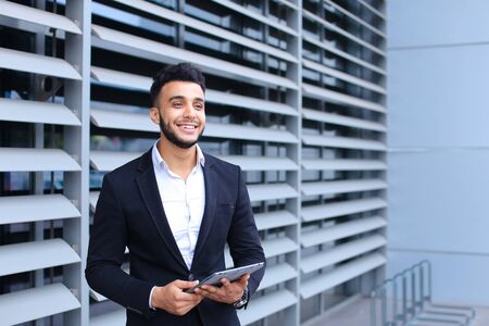 Young handsome businessman Arab Muslims is in the business center in black elegant dress on building background. Businessman man male looking waiting concept and smiling.の写真素材