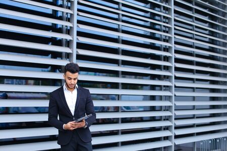 Young handsome businessman Arab Muslims is in the business center in black elegant dress on building background. Businessman man male looking waiting concept and smiling.の写真素材